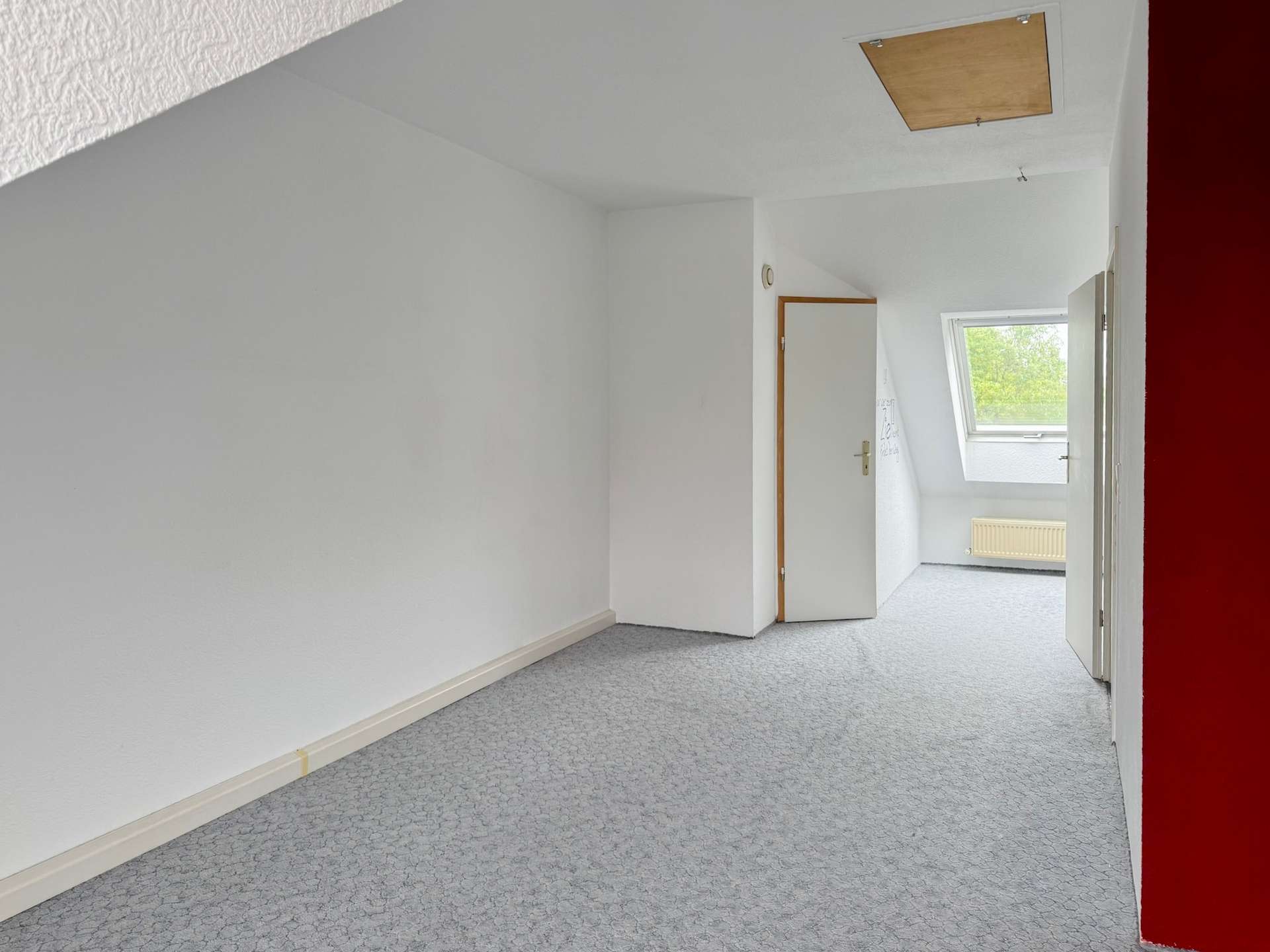 Empty white hallway with a sloped ceiling, open door, and a window letting in daylight; pale patterned carpet and radiator visible.