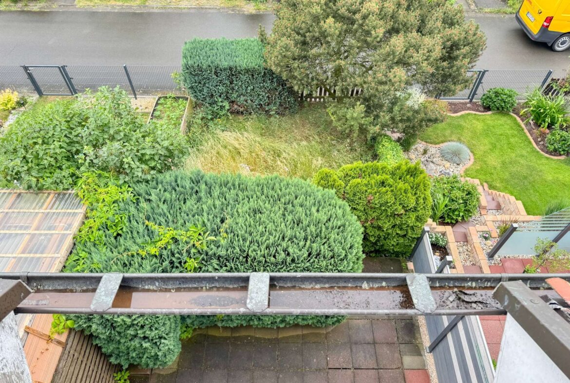 Aerial view of a residential garden with neatly trimmed hedges, a lawn, stone pathways, and a bright yellow van parked on the street beyond a metal fence.