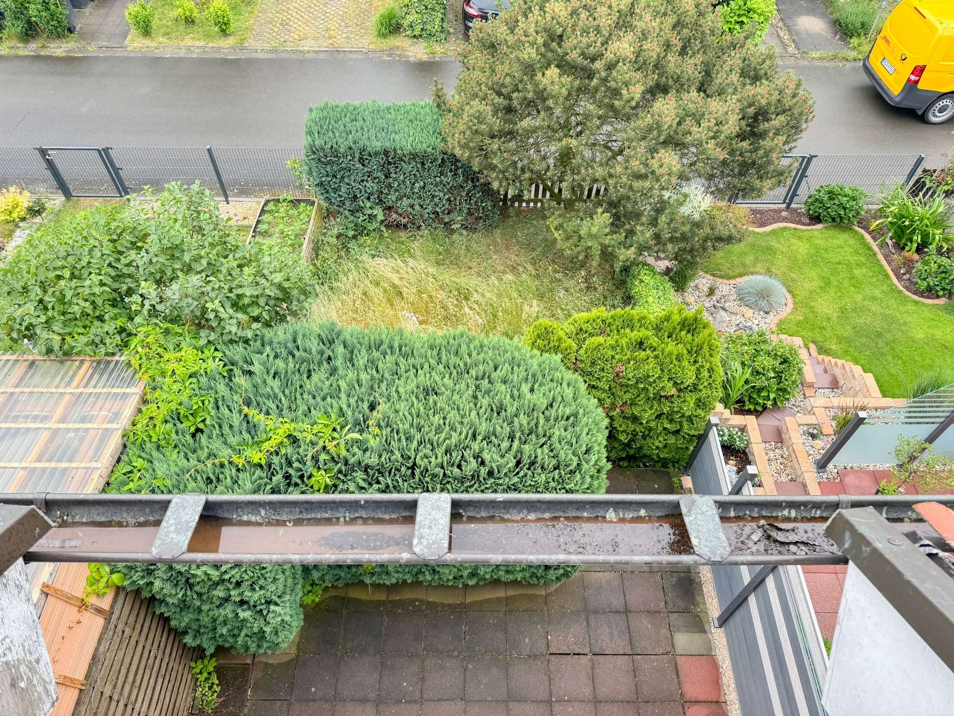 Aerial view of a residential garden with neatly trimmed hedges, a lawn, stone pathways, and a bright yellow van parked on the street beyond a metal fence.