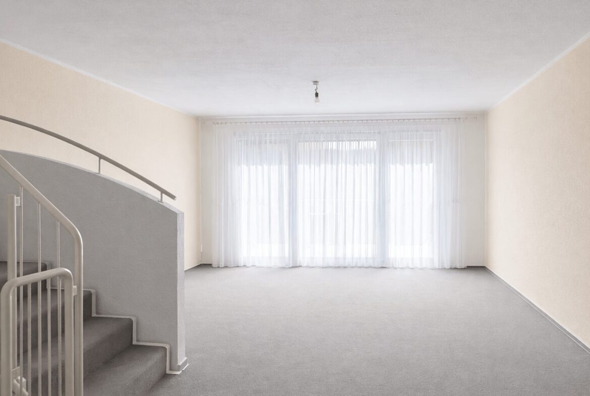 Empty living room with a curved staircase on the left, pale beige walls, and a large window with sheer white curtains letting in light.