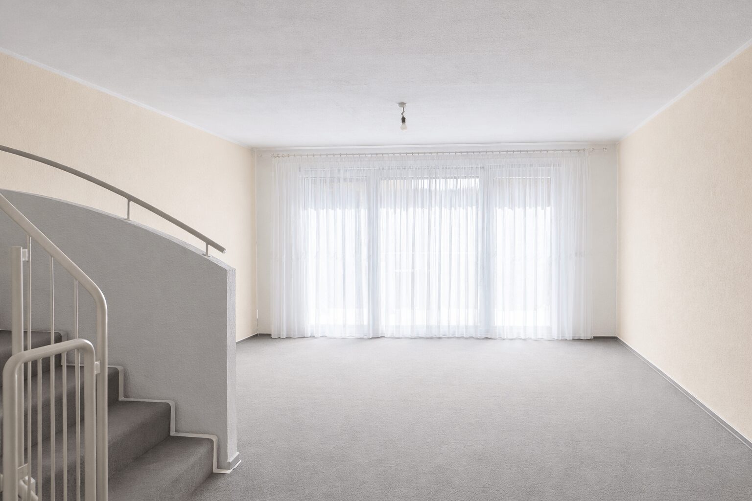 Empty living room with a curved staircase on the left, pale beige walls, and a large window with sheer white curtains letting in light.