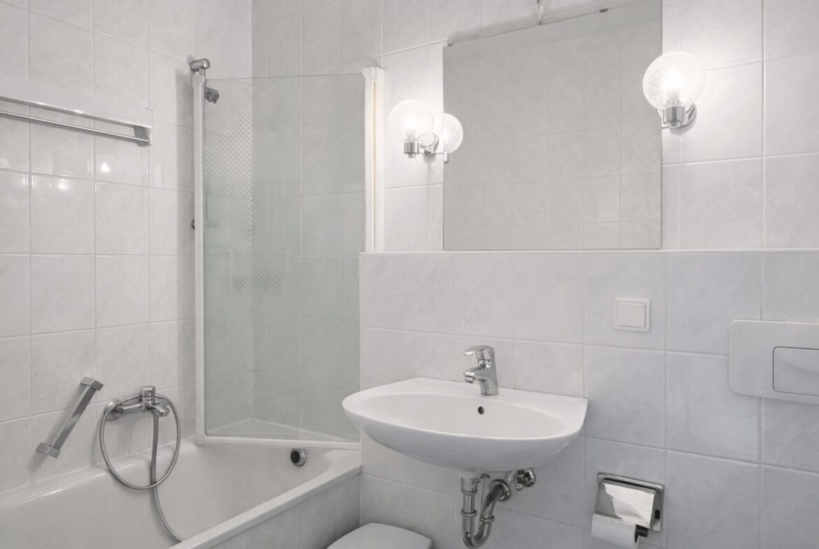 White tiled bathroom with a corner bathtub and glass shower enclosure, a wall-mounted sink, and a large mirror above. A round globe light and chrome fixtures complete the scene.