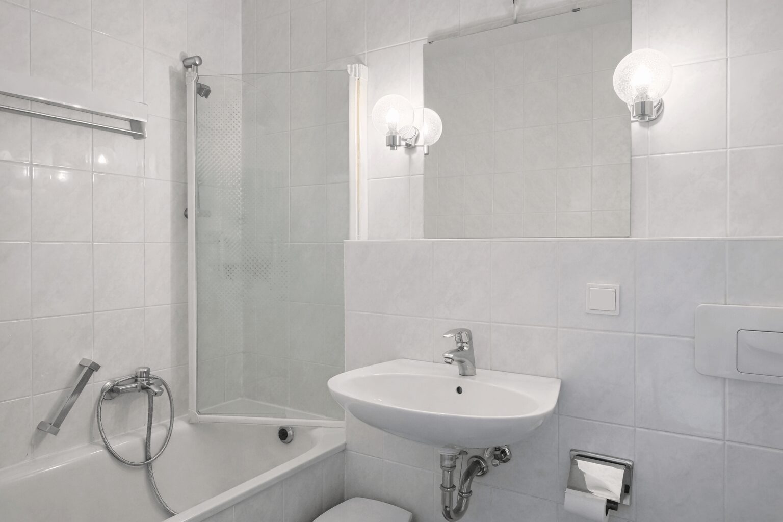 White tiled bathroom with a corner bathtub and glass shower enclosure, a wall-mounted sink, and a large mirror above. A round globe light and chrome fixtures complete the scene.