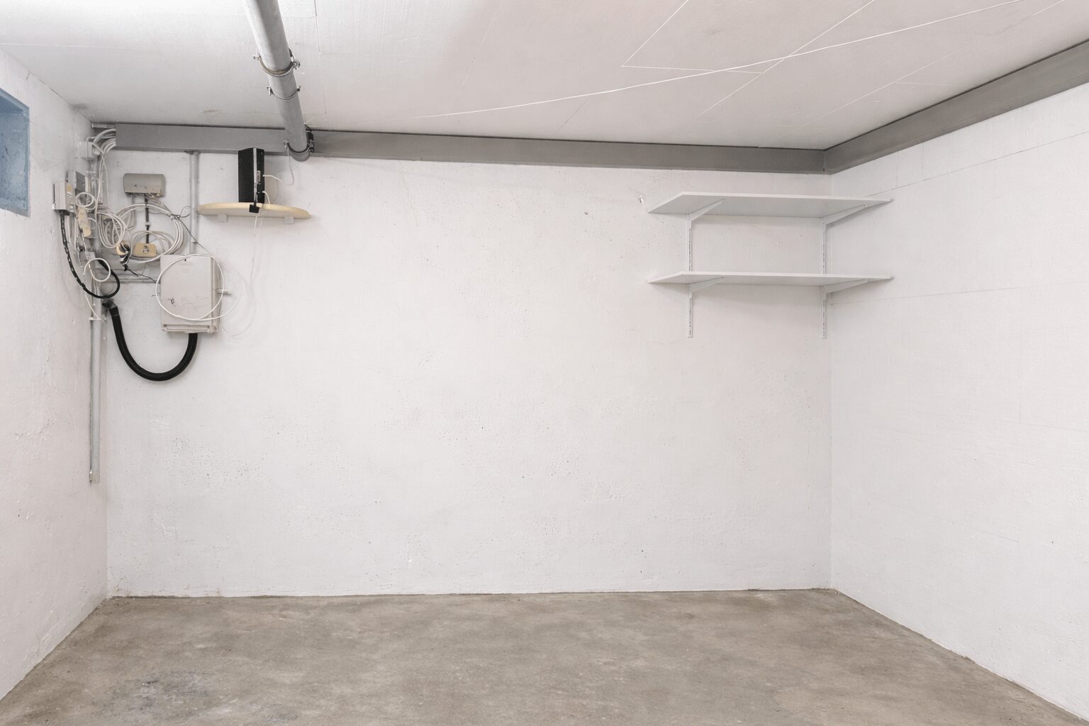 Bare storage room with exposed plumbing and tangled cables on the left, two white shelves on the right wall, concrete floor