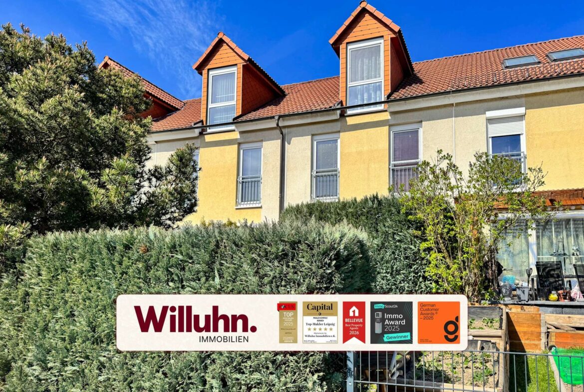 Apartment building with yellow facade, red-tiled roof and two dormer windows behind a dense hedge; a Willuhn Immobilien sign with awards in the foreground.