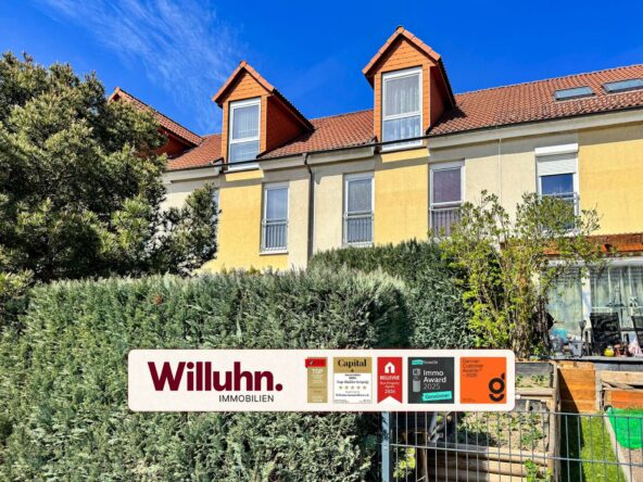Apartment building with yellow facade, red-tiled roof and two dormer windows behind a dense hedge; a Willuhn Immobilien sign with awards in the foreground.