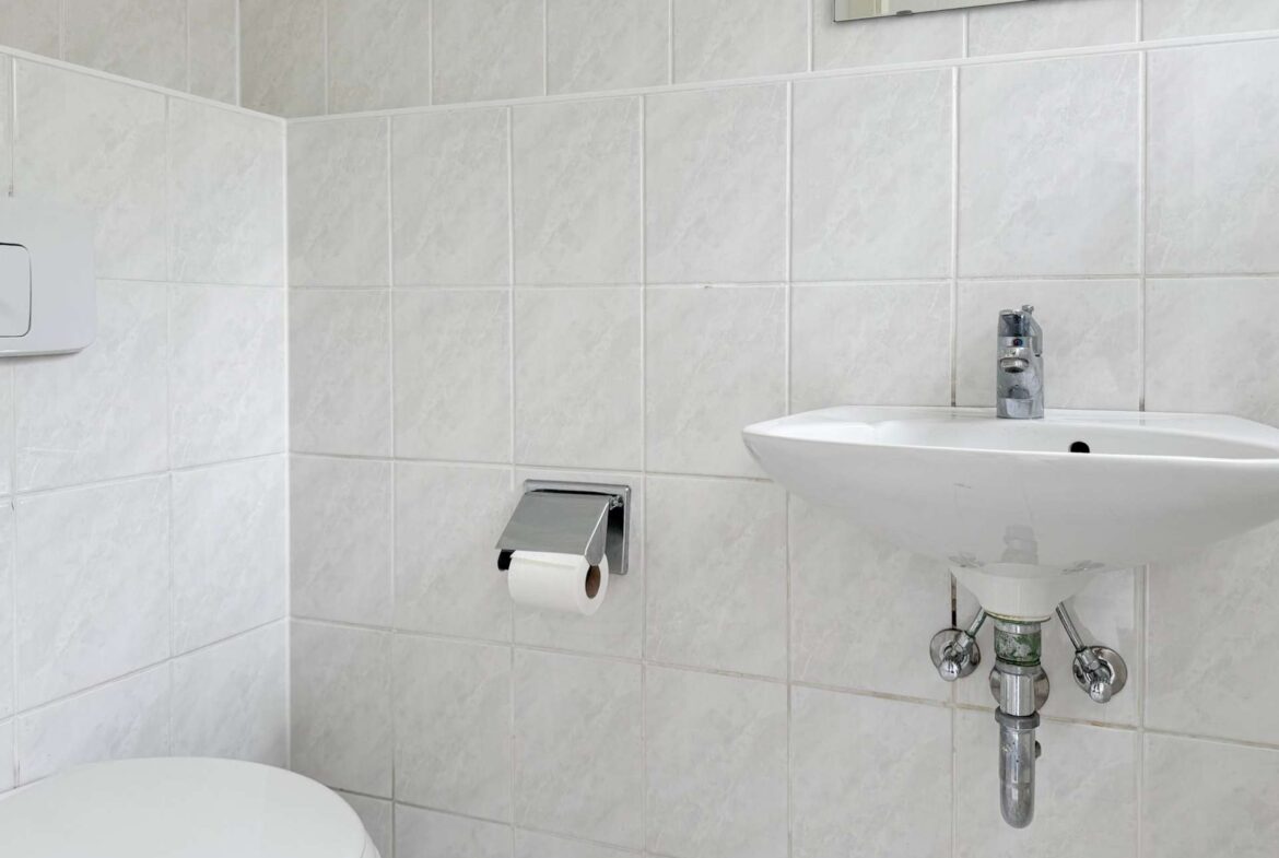 Small bathroom with white tiled walls, a wall-mounted sink with chrome faucet, exposed plumbing, a toilet, and a toilet paper holder.