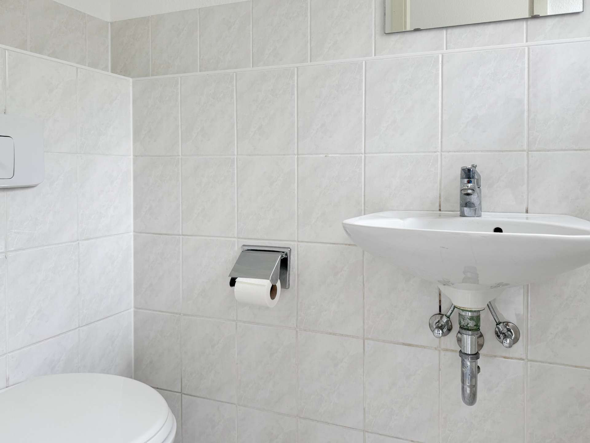 Small bathroom with white tiled walls, a wall-mounted sink with chrome faucet, exposed plumbing, a toilet, and a toilet paper holder.