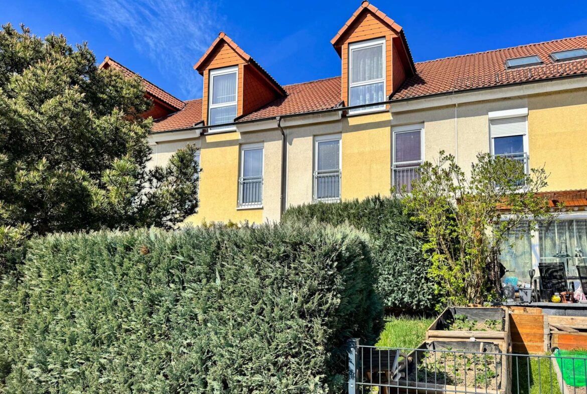 Residential building with yellow walls, red-tiled roof and two dormer windows, framed by dense evergreen hedges under a blue sky.
