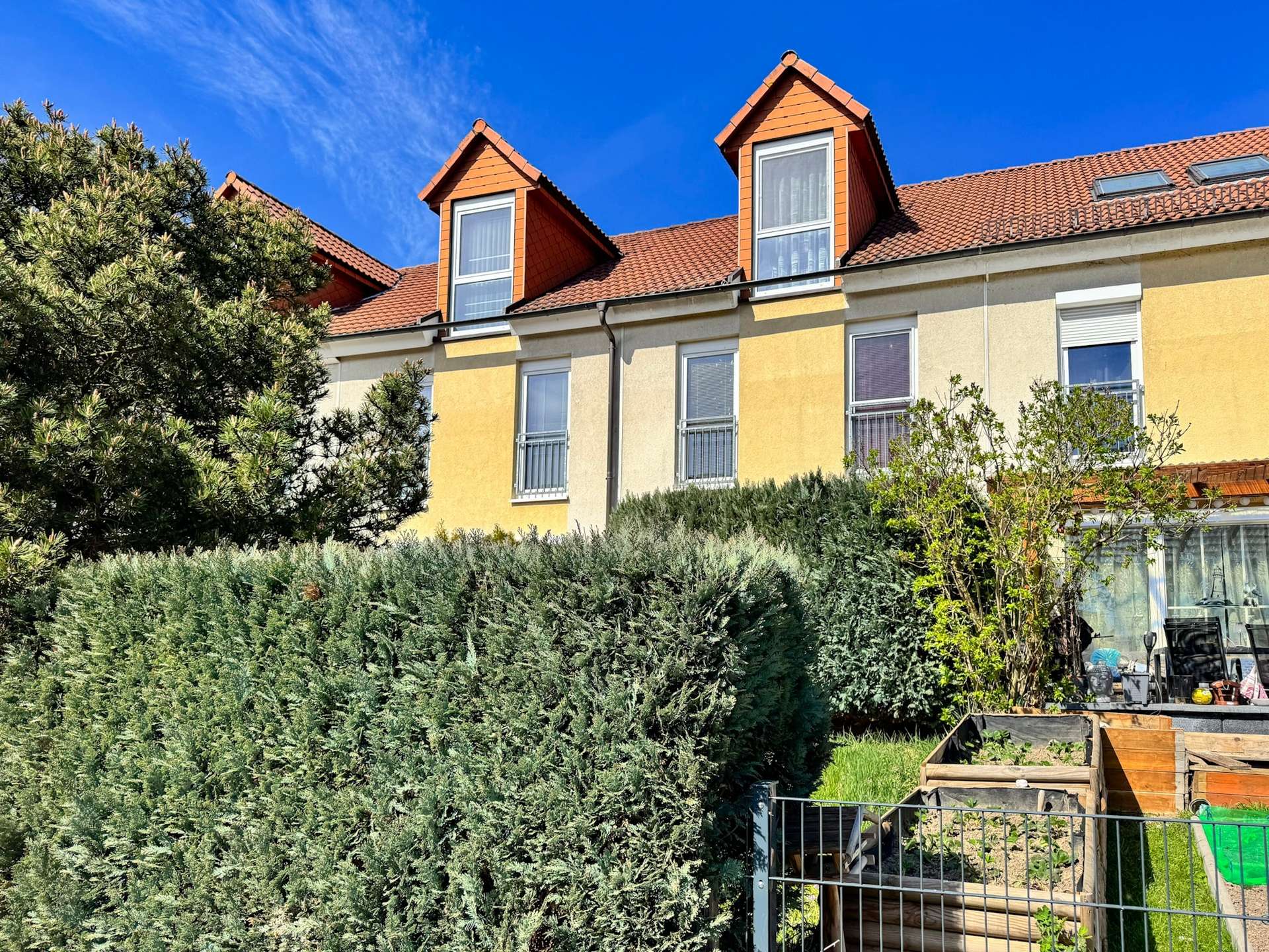 Residential building with yellow walls, red-tiled roof and two dormer windows, framed by dense evergreen hedges under a blue sky.