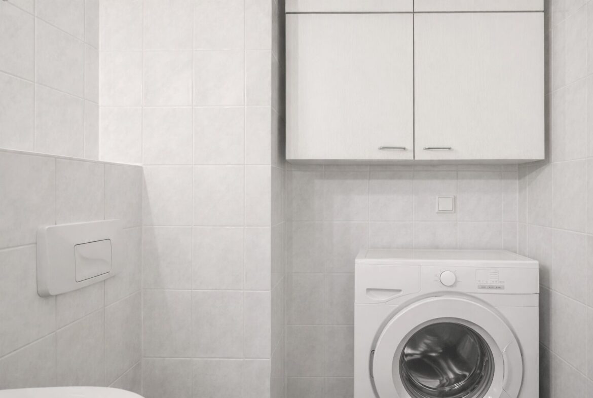 White front-loading washing machine under a wall cabinet in a white-tiled utility bathroom.