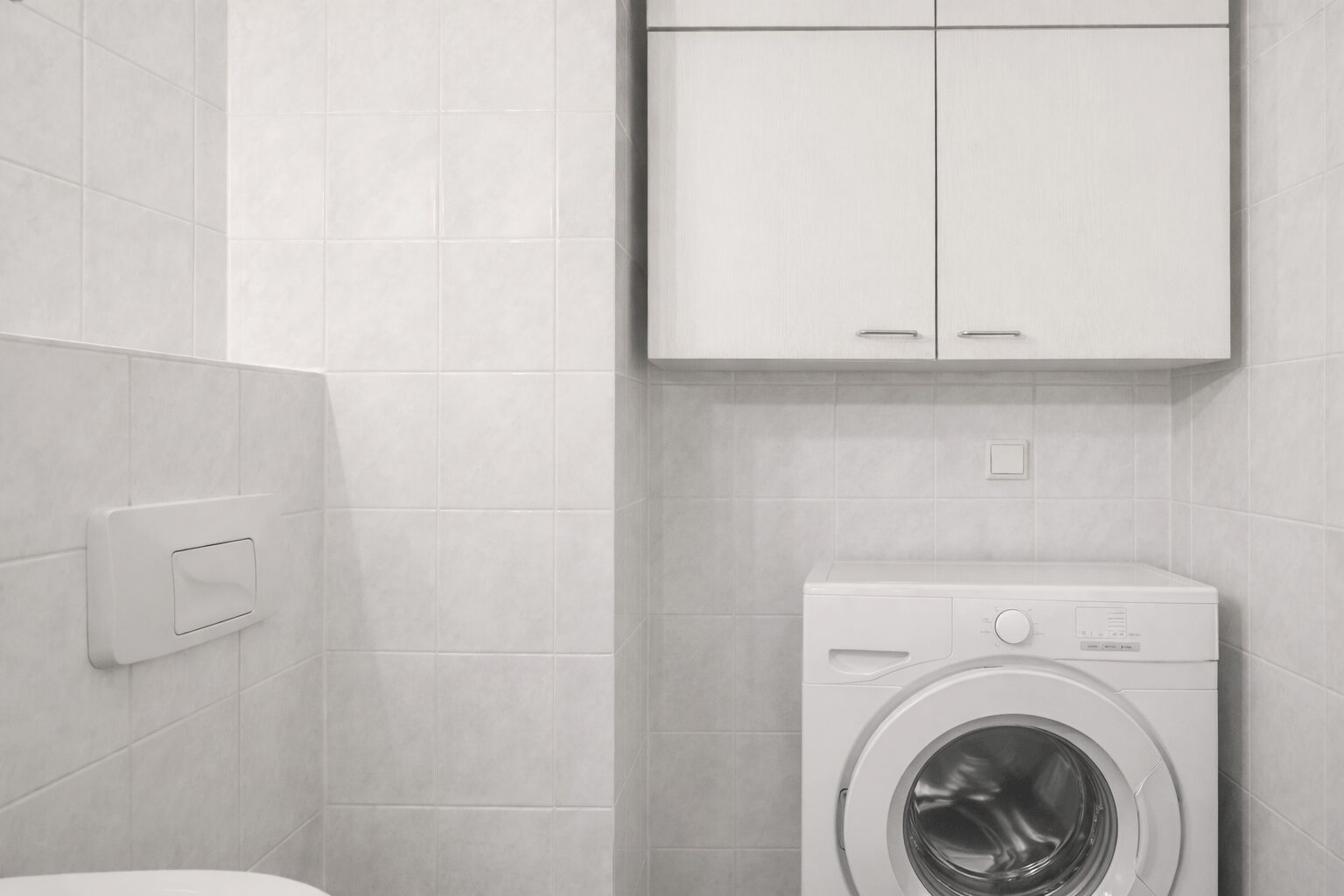 White front-loading washing machine under a wall cabinet in a white-tiled utility bathroom.