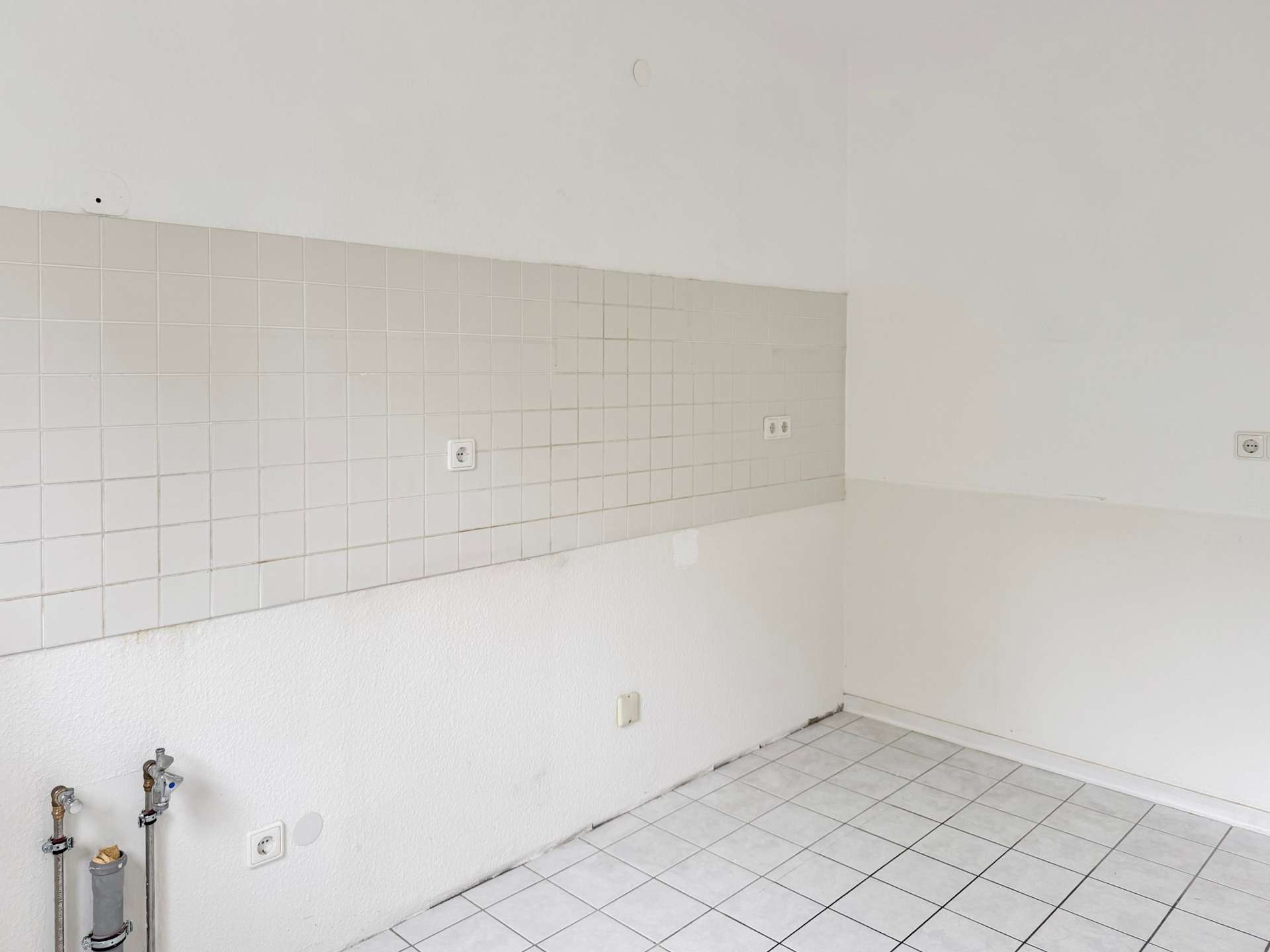 Empty kitchen/utility space with white tiled backsplash, tiled floor, and exposed plumbing on the left wall.