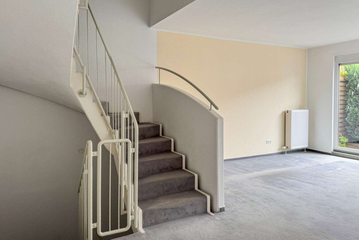 Interior stairwell with a curved handrail, beige accent wall, and large window bringing in natural light, revealing a garden outside.