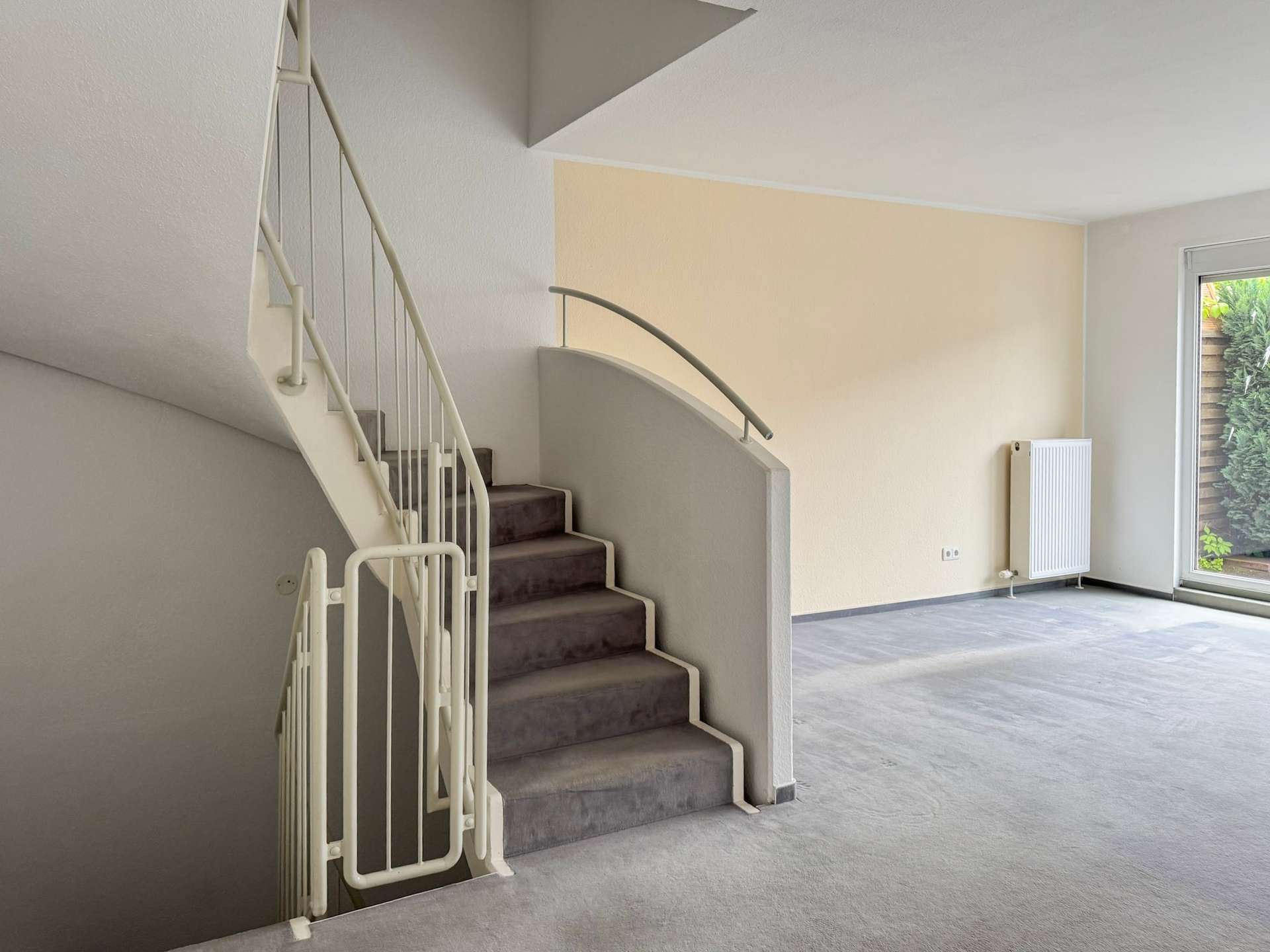 Interior stairwell with a curved handrail, beige accent wall, and large window bringing in natural light, revealing a garden outside.
