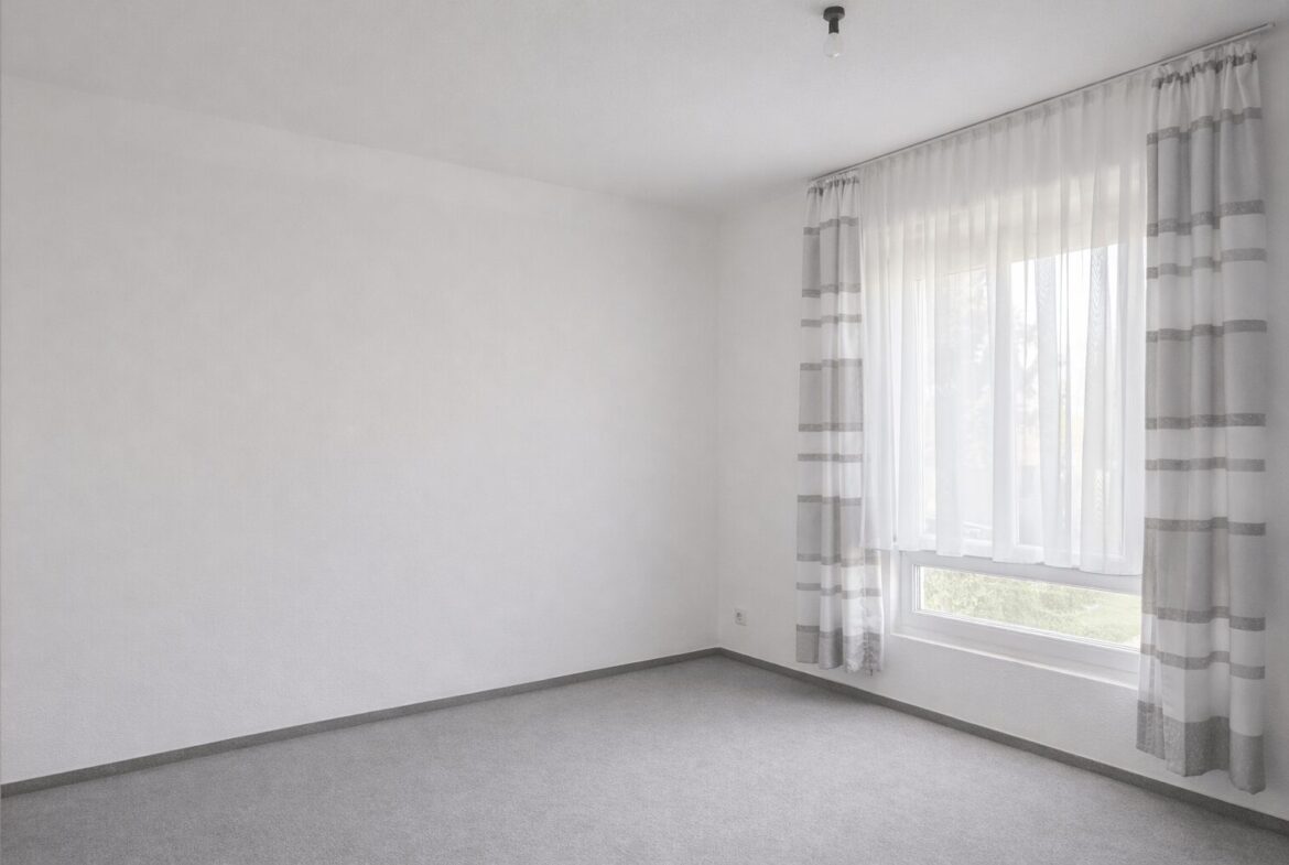Empty bright room with white walls, gray carpet, and a window with sheer white curtains.