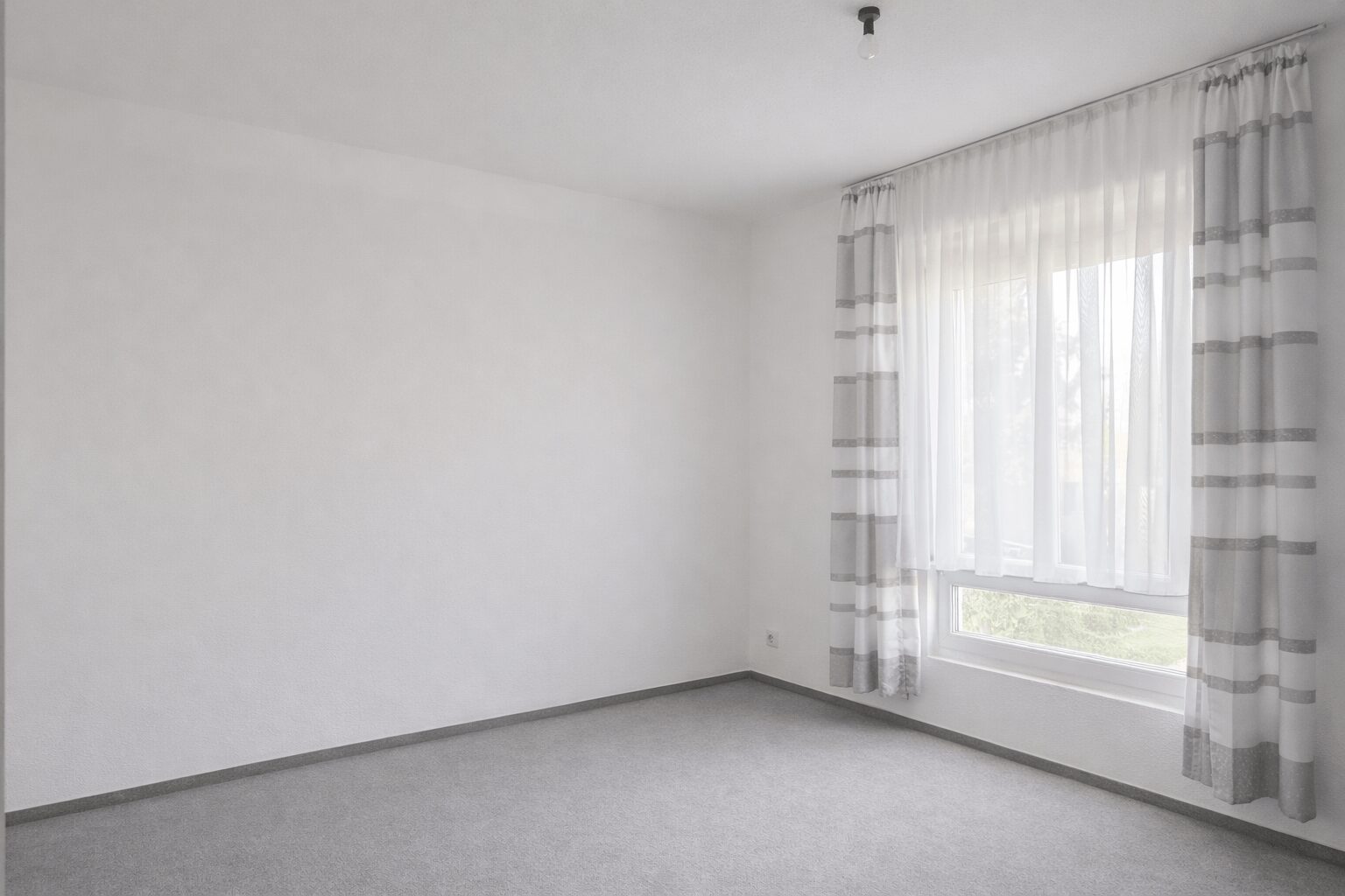Empty bright room with white walls, gray carpet, and a window with sheer white curtains.