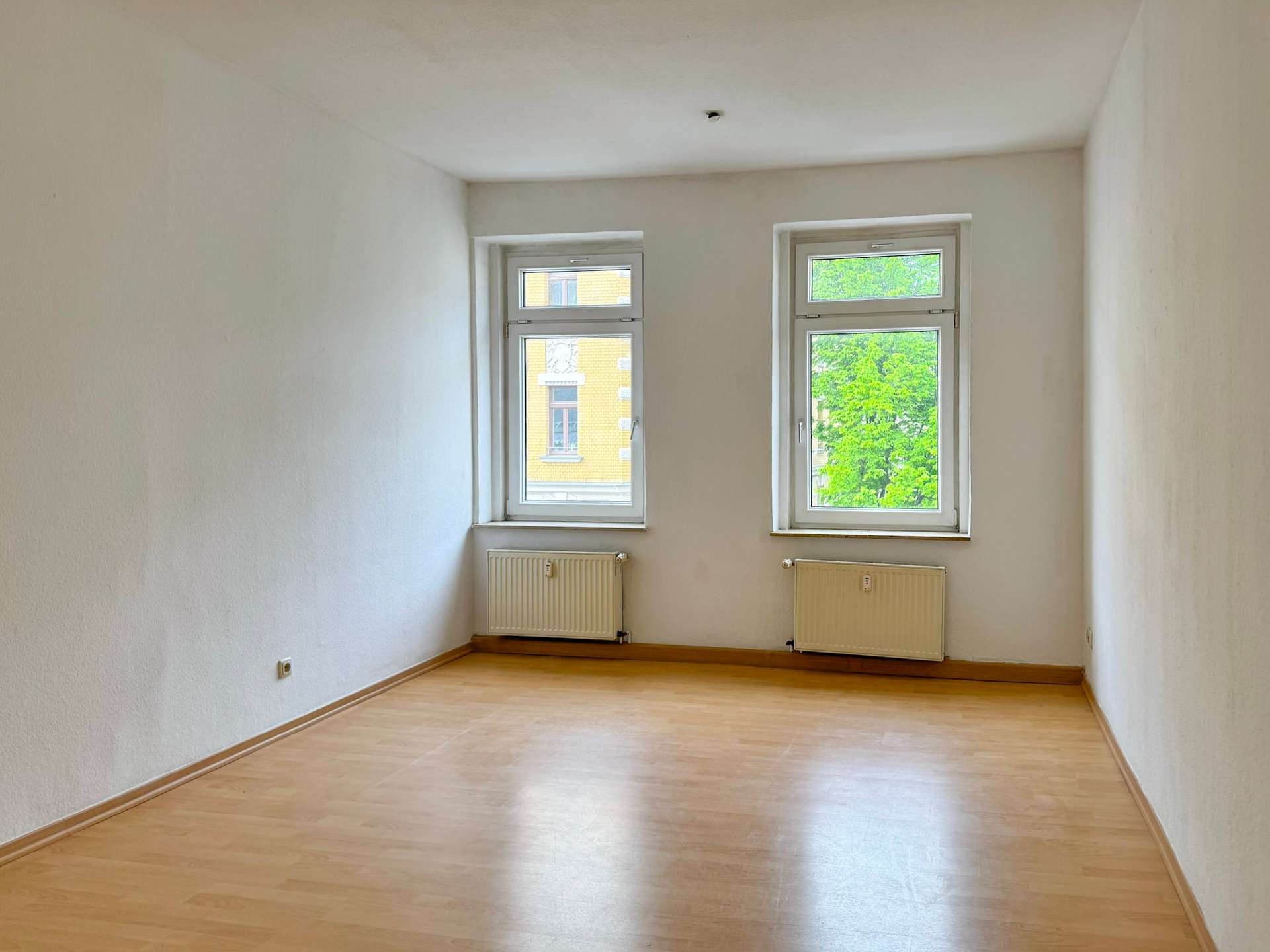 Empty rectangular room with light wood flooring, two windows, and radiators under them against white walls.