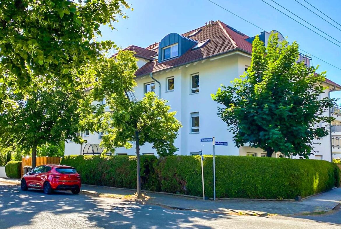 White apartment building with a red-tiled roof at a street corner, bordered by a tall hedge and leafy trees, with a red car parked nearby.