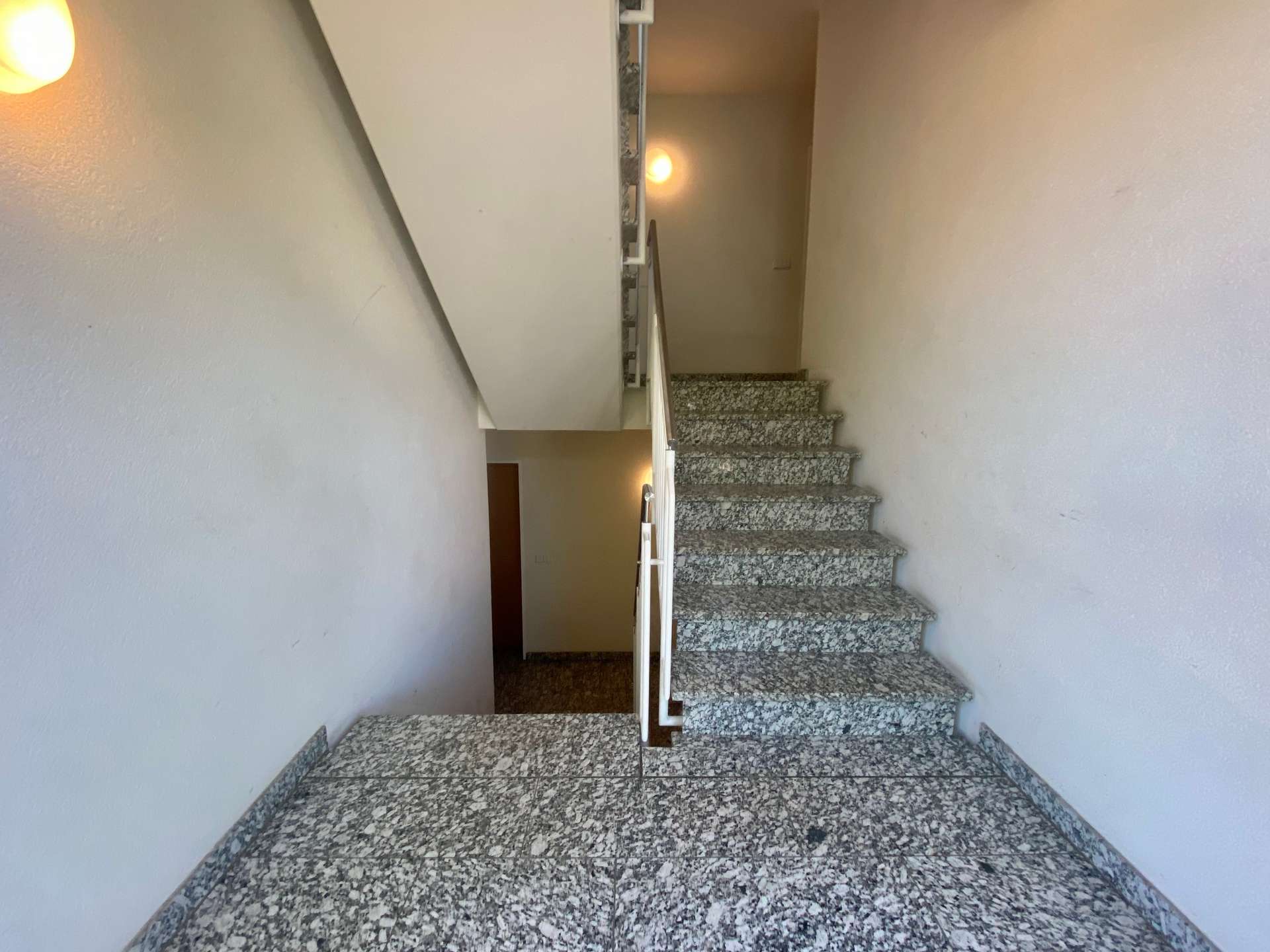 Interior stairwell with terrazzo steps and metal handrail, beige walls, lit by wall lamps.