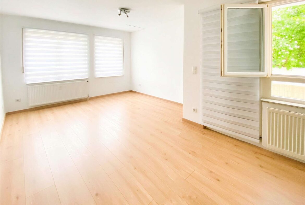 Bright empty living room with light wood floors, white walls, and large windows with horizontal blinds; open window shows greenery outside.