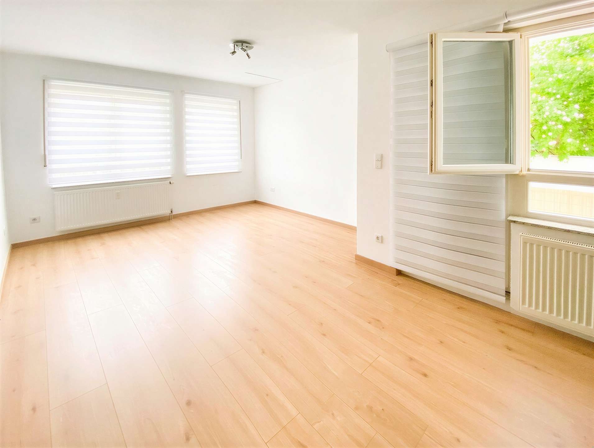 Bright empty living room with light wood floors, white walls, and large windows with horizontal blinds; open window shows greenery outside.