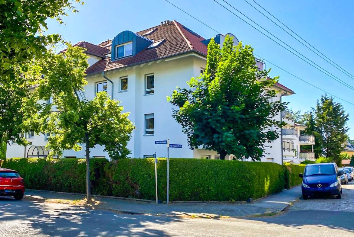 White multi-story apartment building on a tree-lined street with a tall hedge and parked cars nearby.