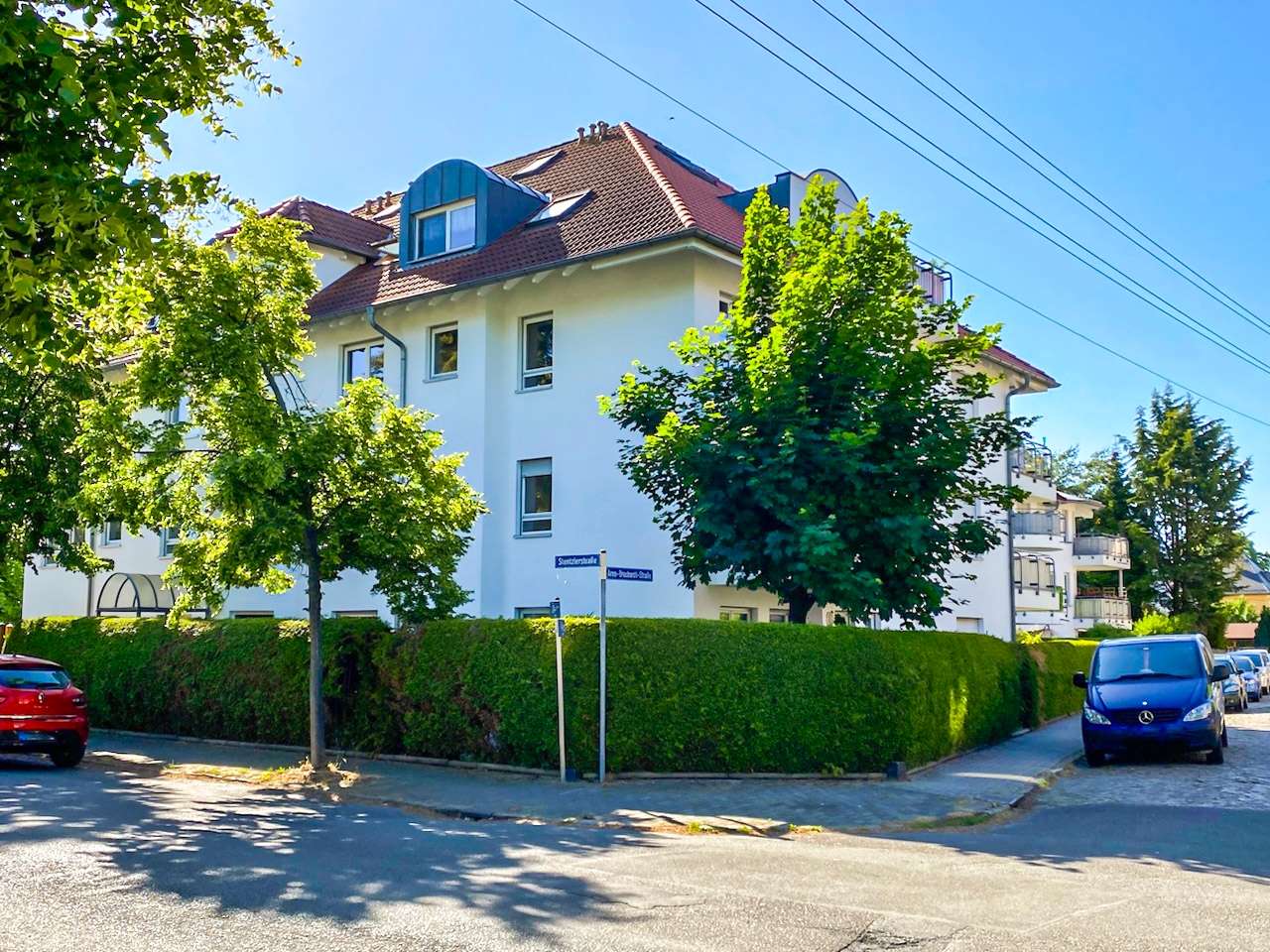 White multi-story apartment building on a tree-lined street with a tall hedge and parked cars nearby.