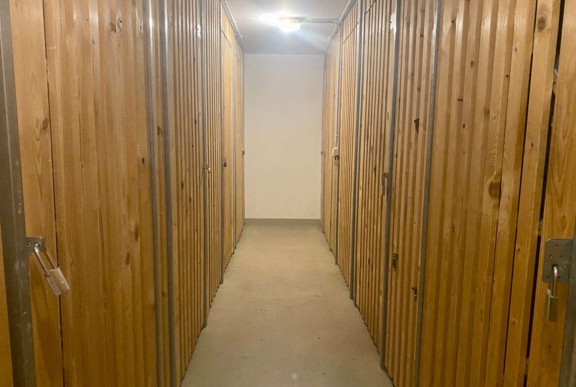 Narrow hallway of wooden storage lockers with metal frames and padlocks, lit by a ceiling light.