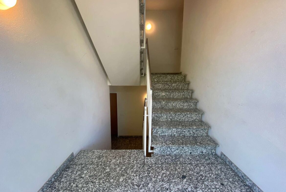 Interior stairwell with terrazzo/granite steps, white walls, and a metal handrail, lit by wall-mounted lamps.