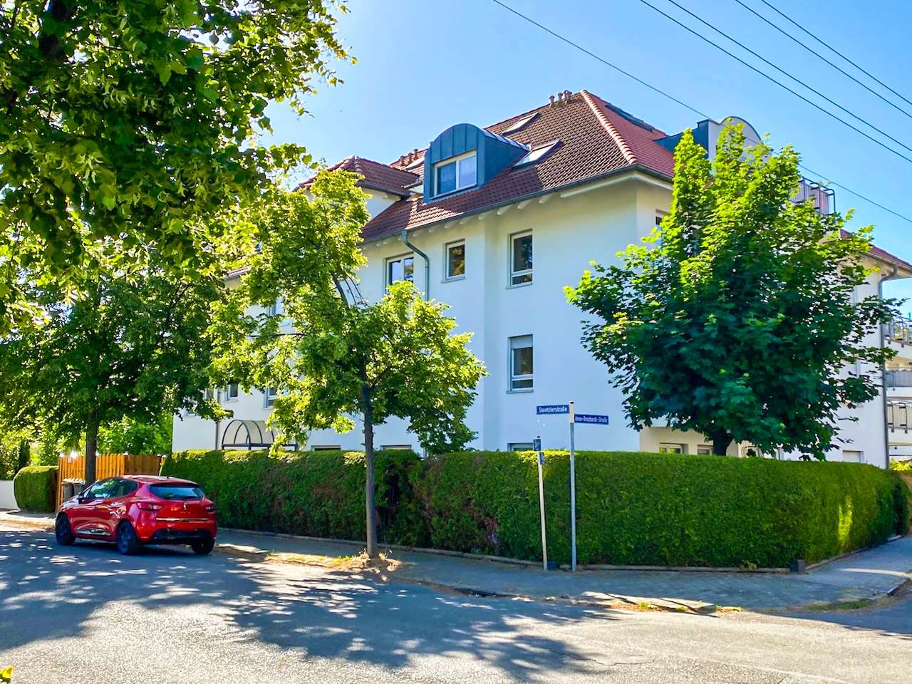 White multi-story residential building with red-tiled roof on a sunlit street, green hedges and trees lining the sidewalk, a red car parked nearby.