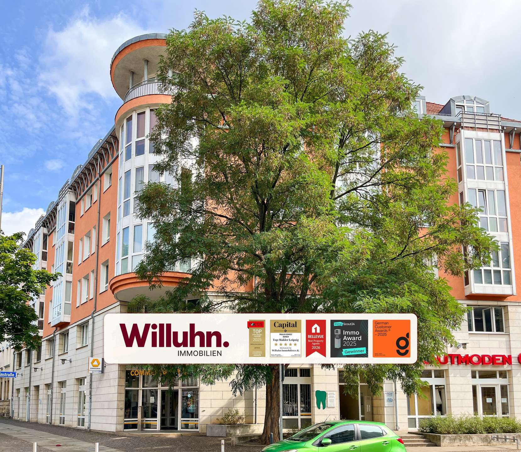 Orange multi-story building with large glass windows partially obscured by a green tree; storefront sign 'Willuhn Immobilien'.