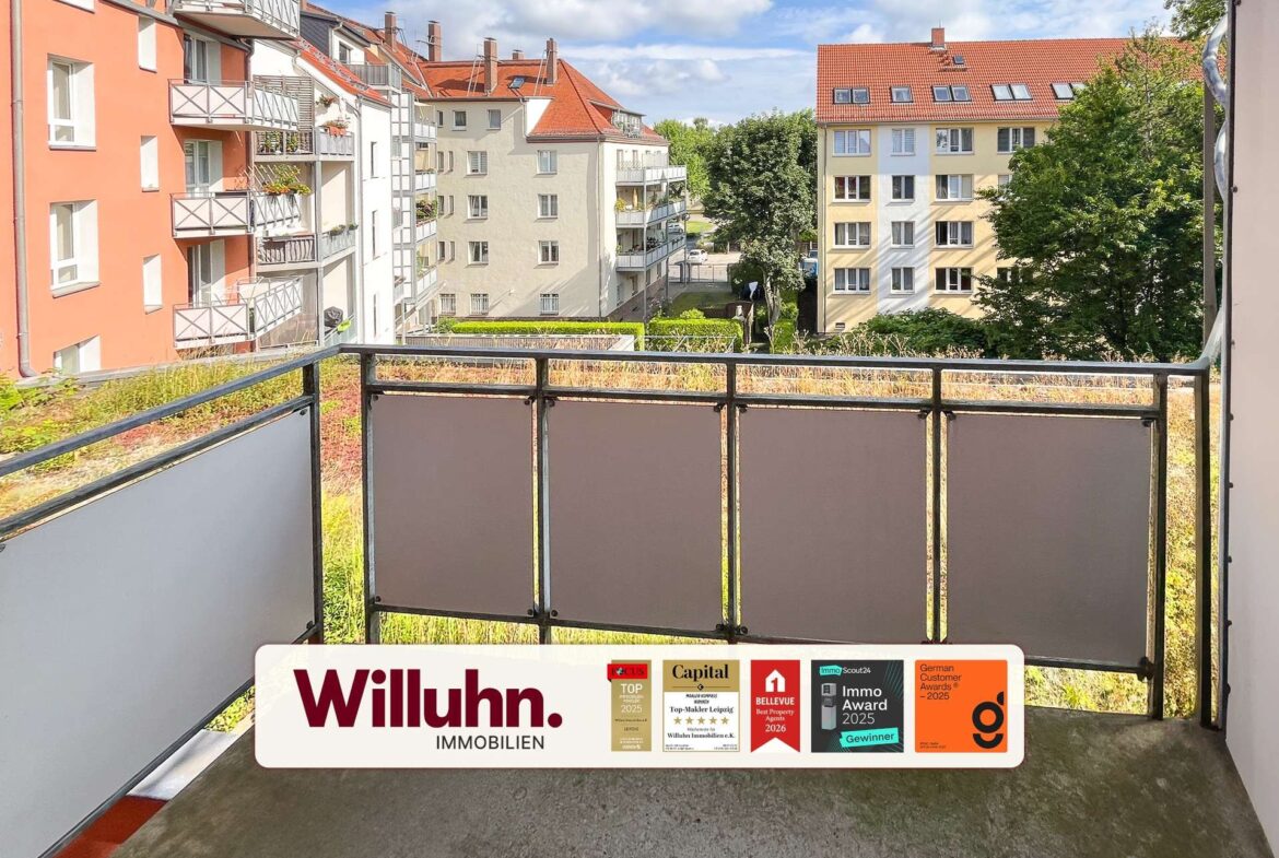 Balcony railing in foreground with red-roofed apartment buildings and trees beyond; a real estate sign 'Willuhn Immobilien' across the bottom.