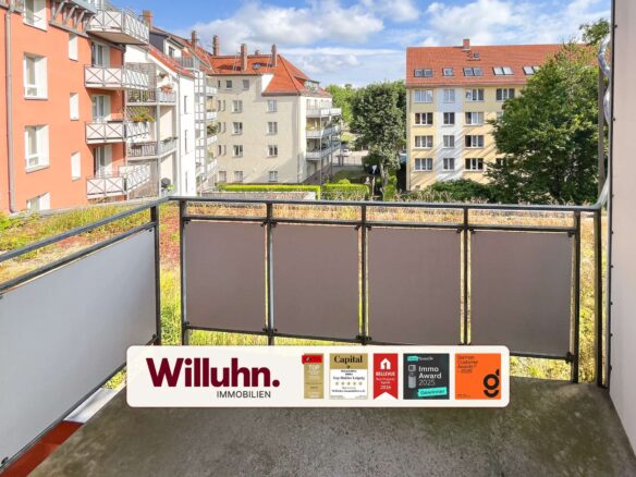 Balcony railing in foreground with red-roofed apartment buildings and trees beyond; a real estate sign 'Willuhn Immobilien' across the bottom.