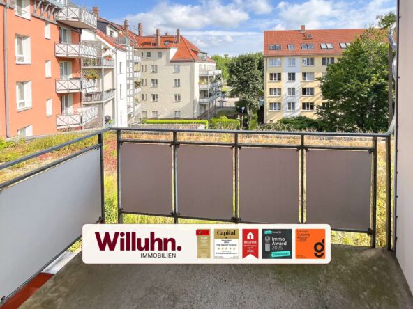 Balcony railing in foreground with red-roofed apartment buildings and trees beyond; a real estate sign 'Willuhn Immobilien' across the bottom.