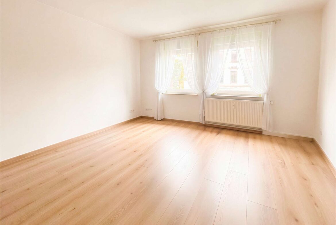 Bright empty room with pale walls, light wood flooring, and a large window dressed in sheer white curtains letting in sunlight, with a radiator beneath the window.