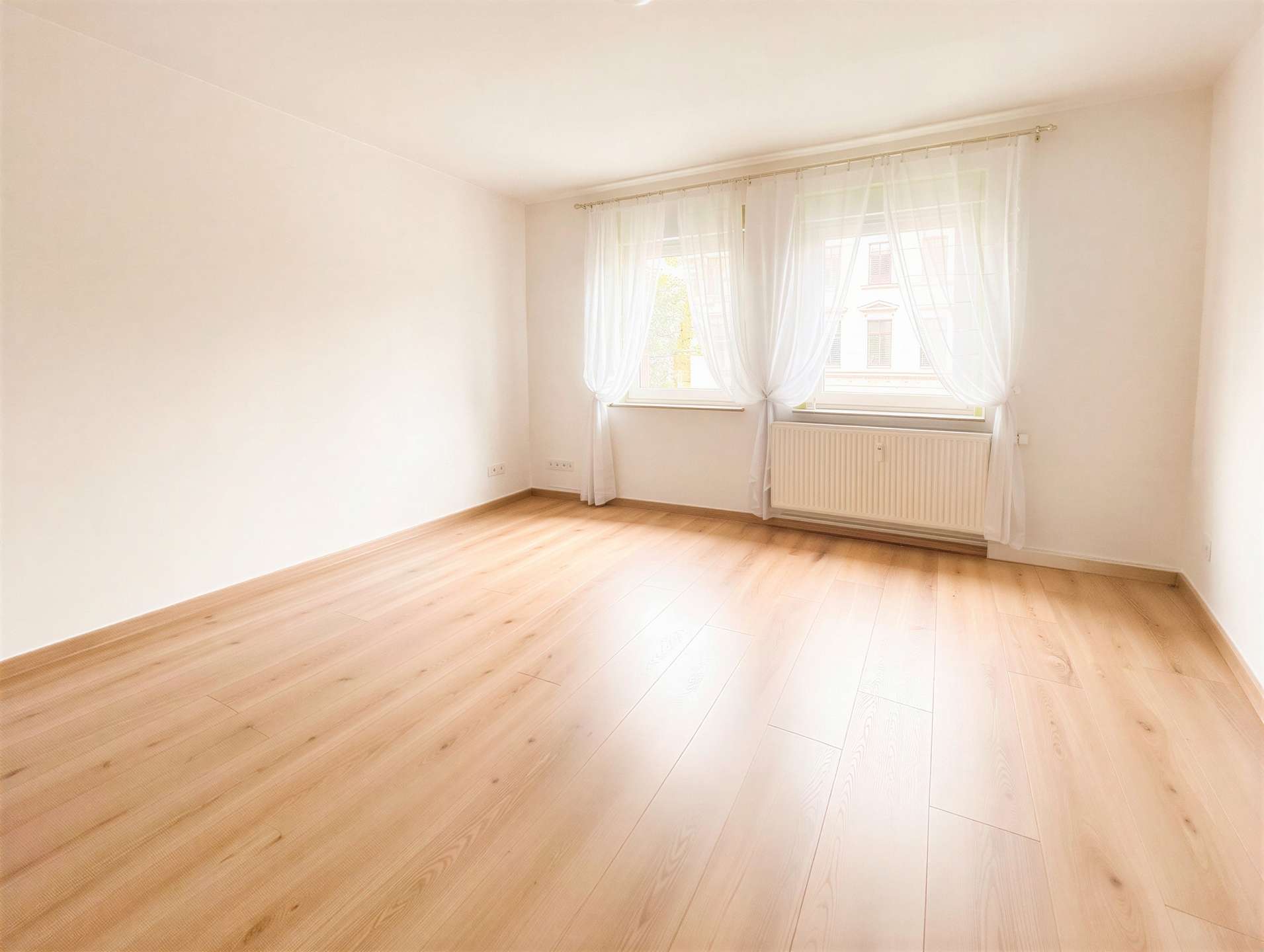 Bright empty room with pale walls, light wood flooring, and a large window dressed in sheer white curtains letting in sunlight, with a radiator beneath the window.