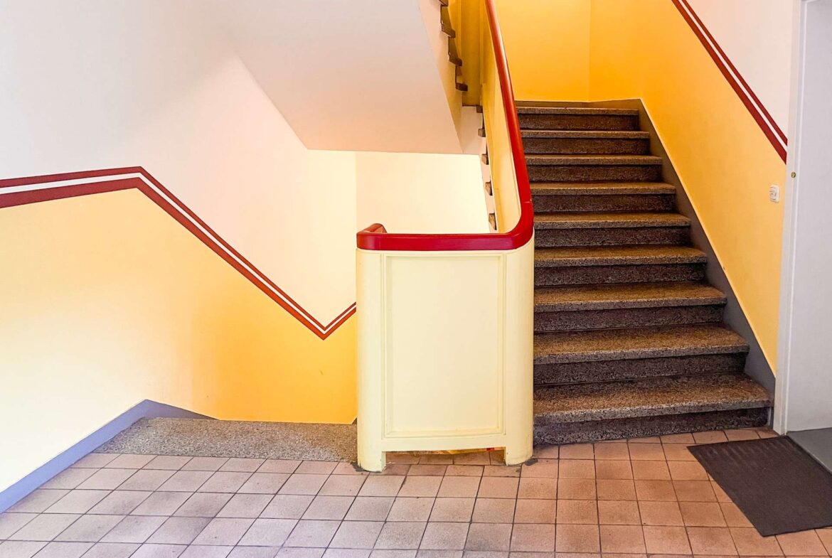 Staircase in a building with yellow walls and red trim, dark steps, and tiled floor area at the bottom left.