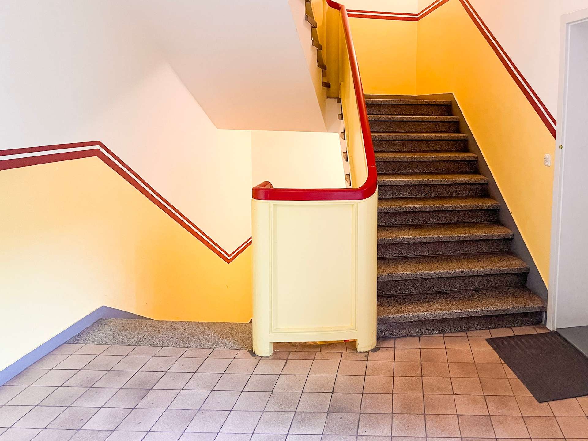 Staircase in a building with yellow walls and red trim, dark steps, and tiled floor area at the bottom left.