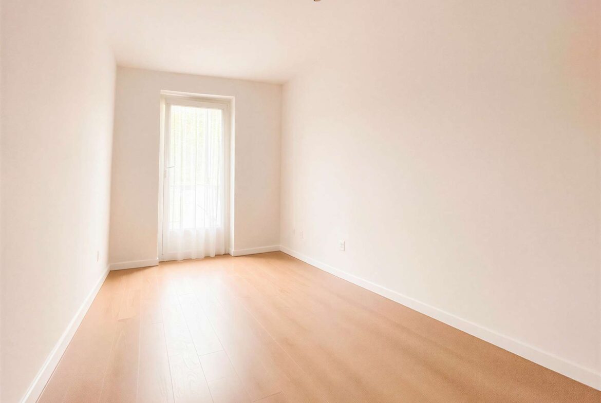 Empty bright room with pale walls, light wood floor, and a window at the far end letting in daylight.