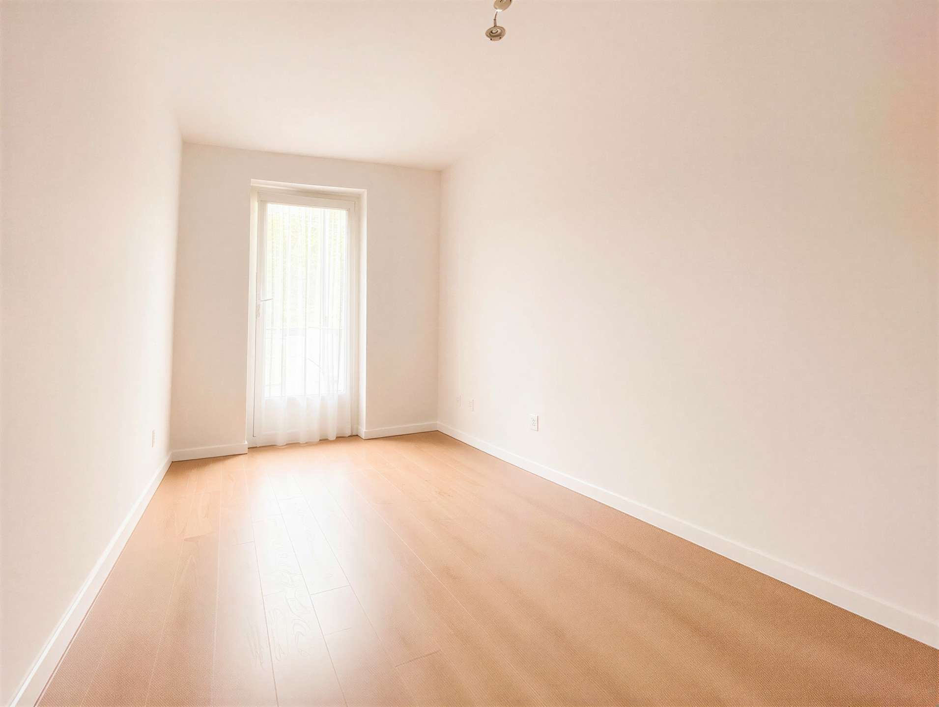 Empty bright room with pale walls, light wood floor, and a window at the far end letting in daylight.
