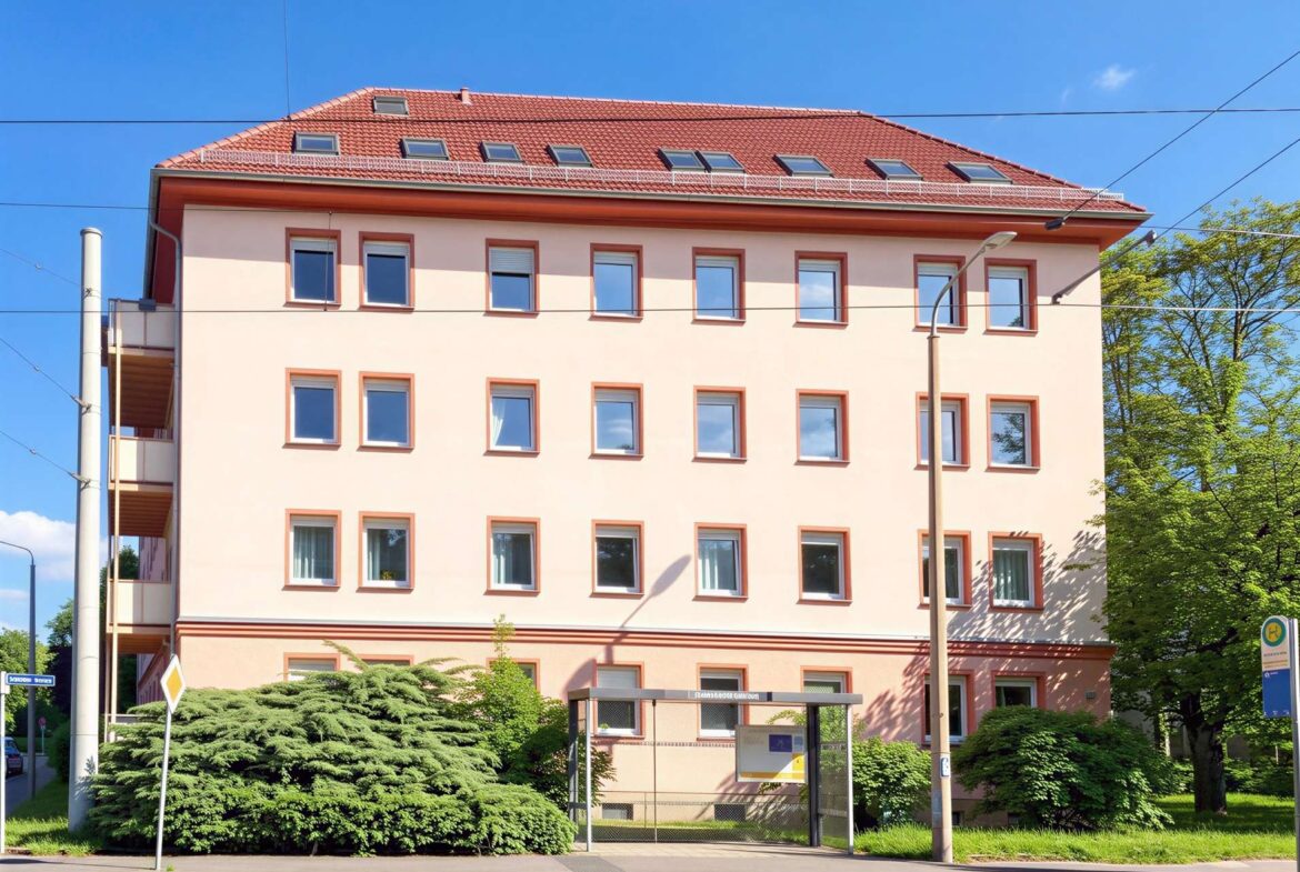Tall peach-colored apartment building with red-tiled roof, multiple evenly spaced windows, under blue sky with overhead tram lines and greenery nearby.