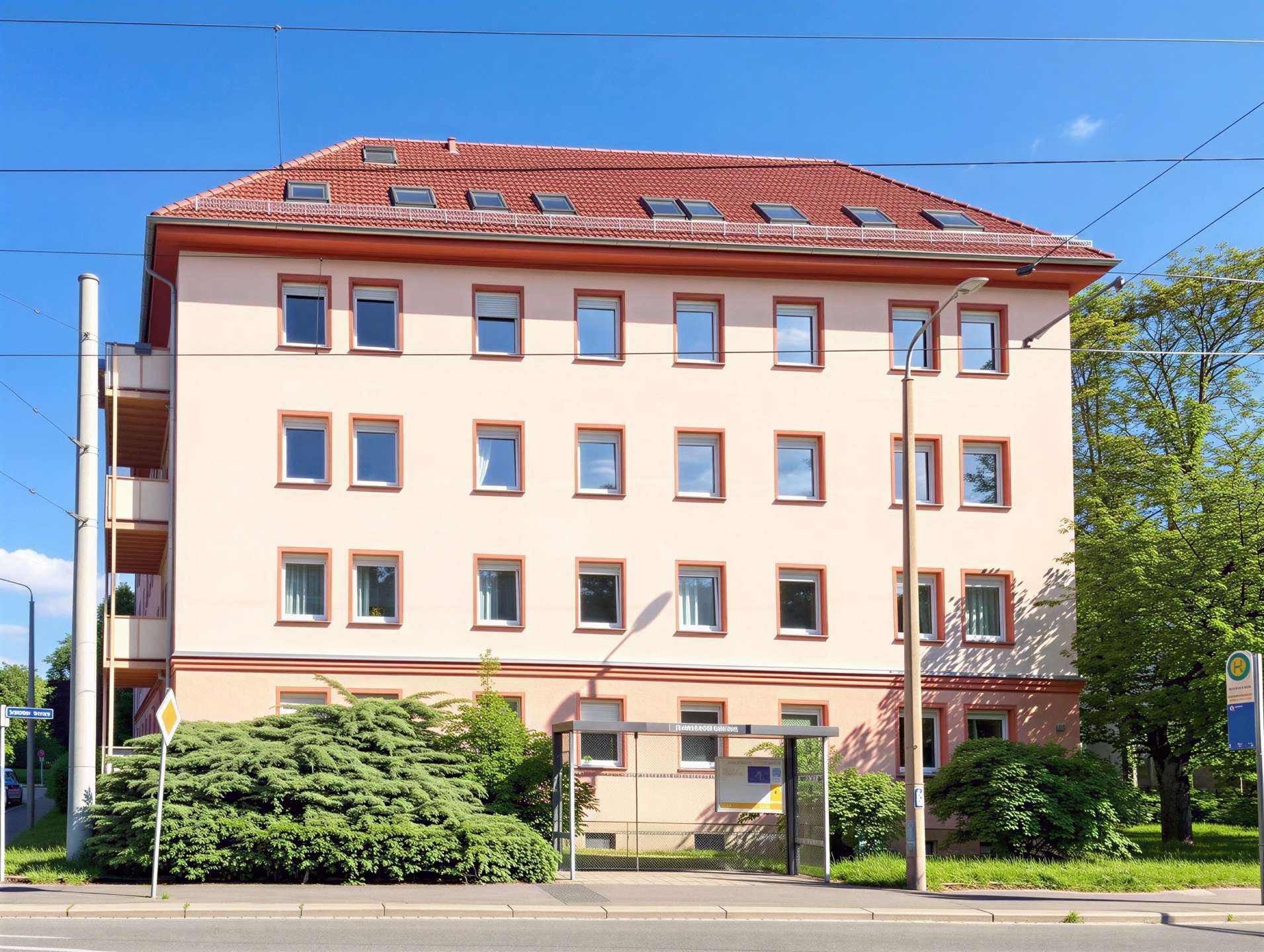 Tall peach-colored apartment building with red-tiled roof, multiple evenly spaced windows, under blue sky with overhead tram lines and greenery nearby.