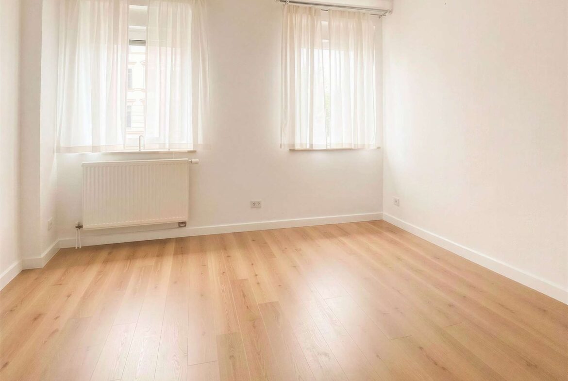 Empty bright room with two large windows covered by sheer white curtains, radiator under the left window, and light wooden flooring.