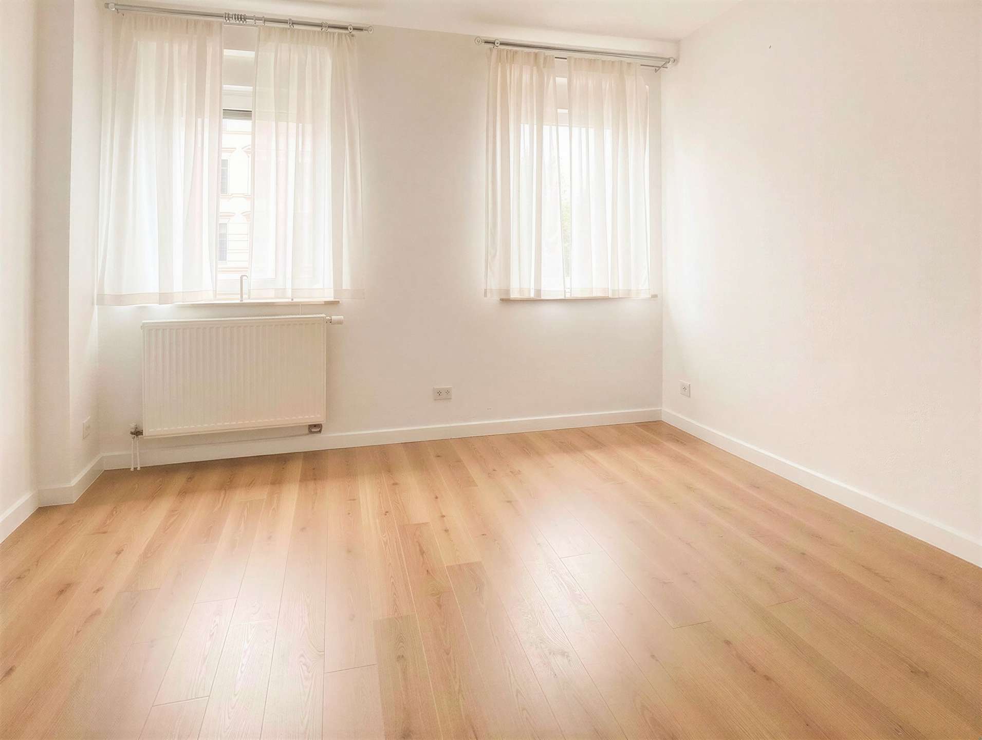 Empty bright room with two large windows covered by sheer white curtains, radiator under the left window, and light wooden flooring.