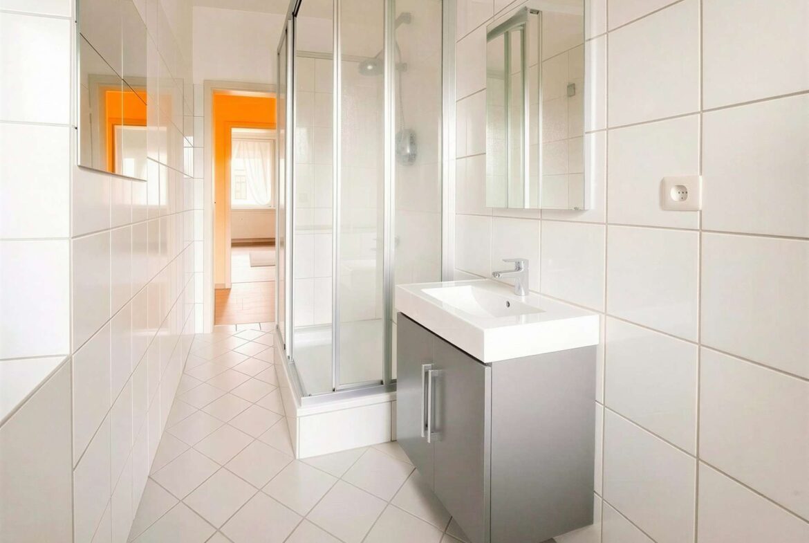 Modern white-tiled bathroom with a glass shower enclosure and a gray vanity; an orange doorway visible in the adjacent room.