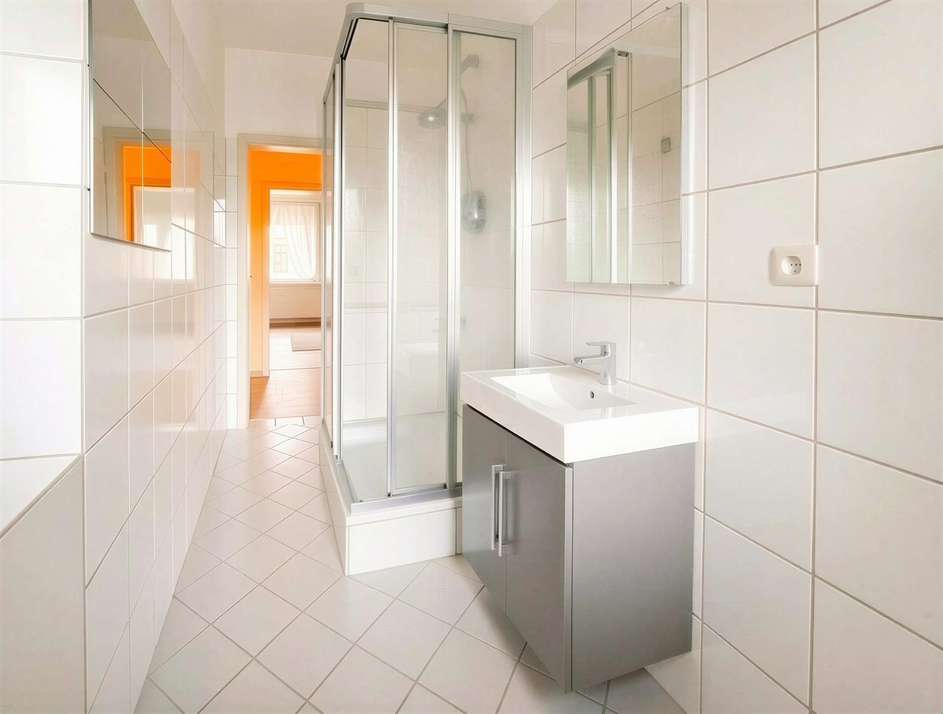 Modern white-tiled bathroom with a glass shower enclosure and a gray vanity; an orange doorway visible in the adjacent room.
