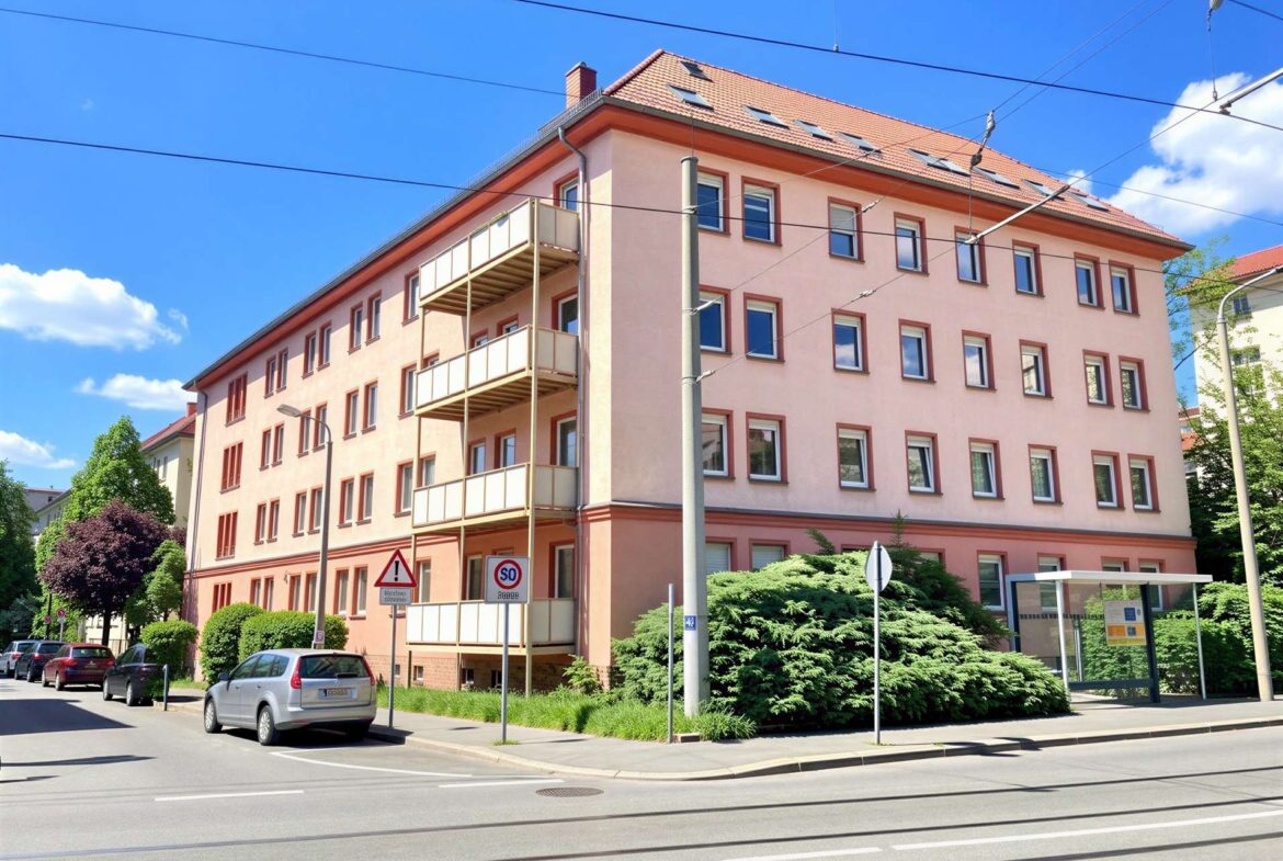Pink apartment building with a red tile roof, balconies on the left, and parked cars along a sunny street.