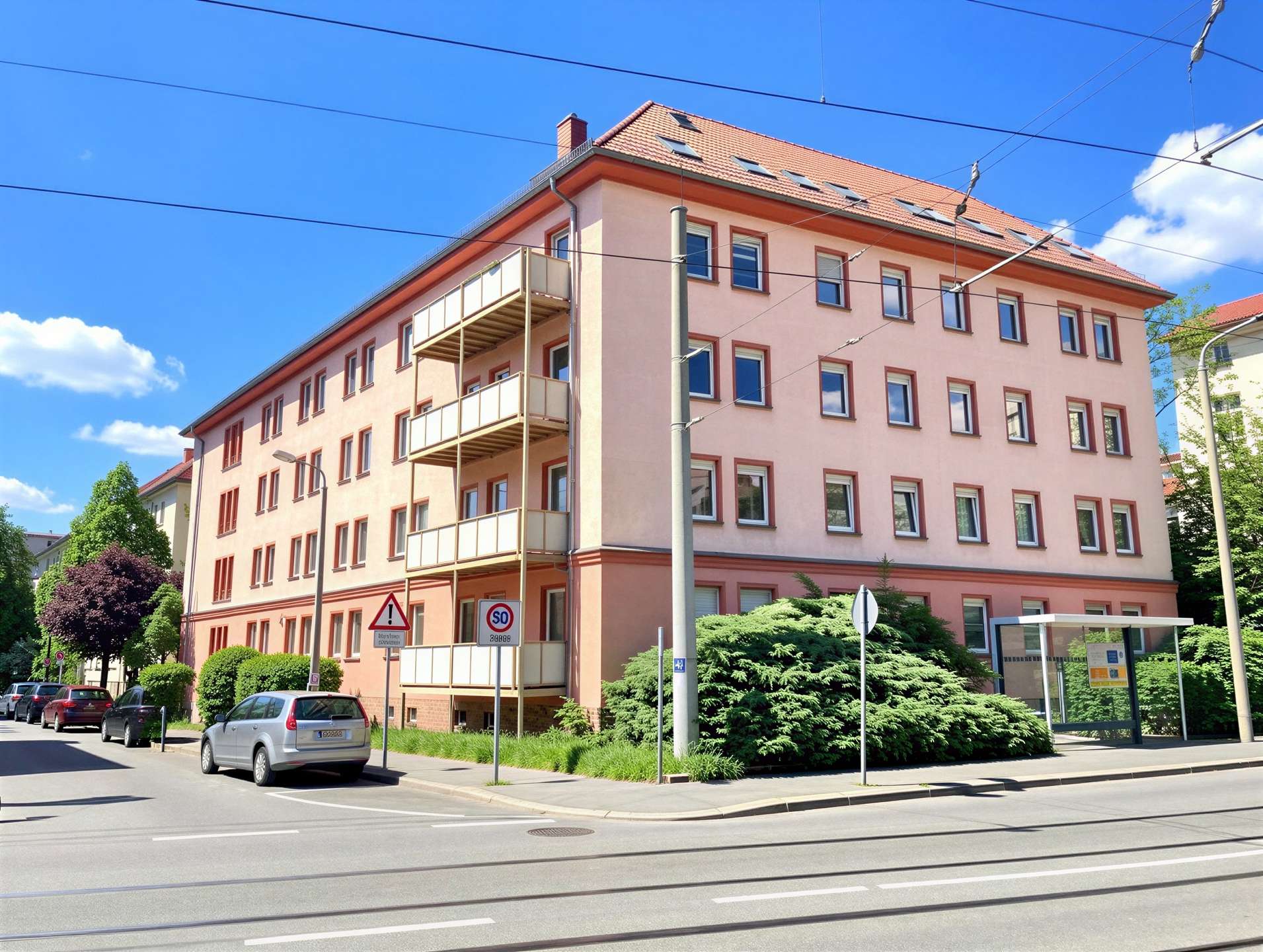 Pink apartment building with a red tile roof, balconies on the left, and parked cars along a sunny street.