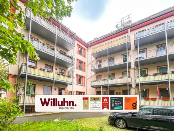 Residential apartment building with metal balconies and greenery, featuring a ground-level Willuhn Immobilien banner.